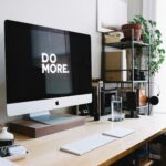 A modern and organized programmers workspace featuring an Apple iMac with "DO MORE." displayed on the screen. A pair of black headphones hangs on a shelf, adding to the sleek and productive setup.