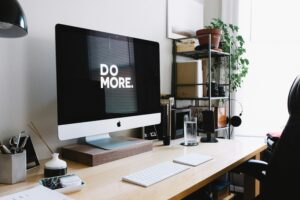 A modern and organized programmers workspace featuring an Apple iMac with "DO MORE." displayed on the screen. A pair of black headphones hangs on a shelf, adding to the sleek and productive setup.